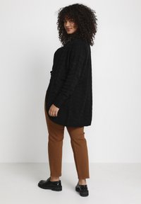 Schwarze Strickjacke mit gerippter Textur, oversized Passform, kombiniert mit braunen Hosen und schwarzen Loafers. Modell sieht nach links, mit lockigem Haar.