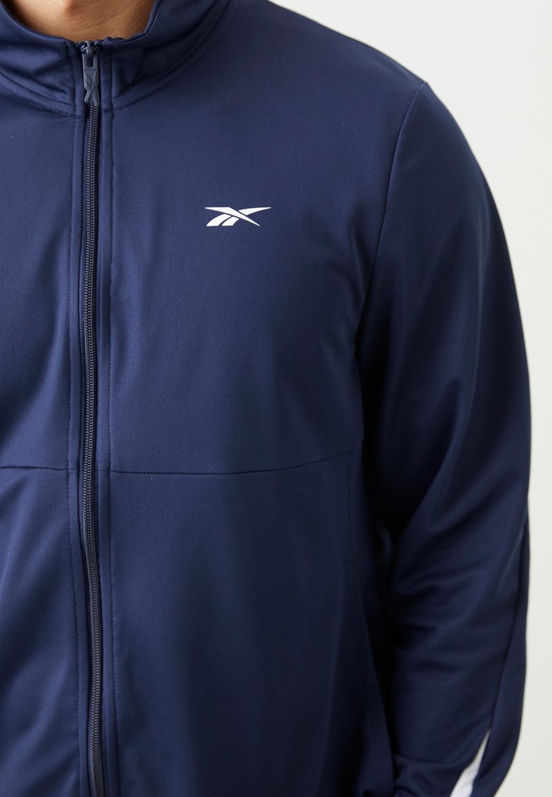 Chaqueta de navy con cremallera y cuello alto, hecha de un tejido suave. Cuenta con un logo blanco en la parte superior izquierda y un detalle de costura horizontal.