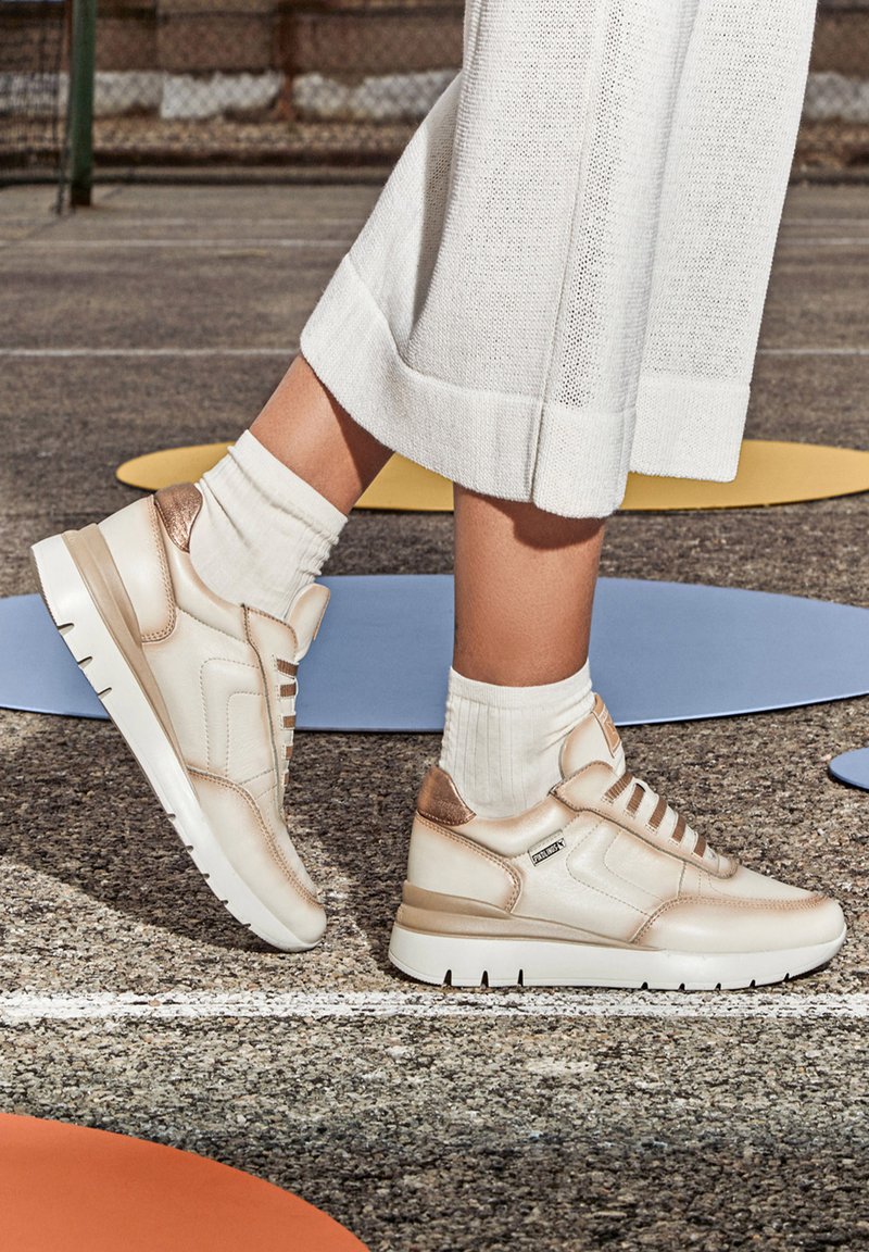 Des baskets beiges avec des accents dorés métalliques, en cuir lisse, des lacets blancs et une semelle texturée, assorties à des chaussettes blanches à côtes.