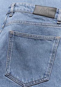 Un jean en denim bleu clair présente une poche arrière avec des coutures beige et une étiquette en cuir noir rectangulaire embossée d'un logo "M".