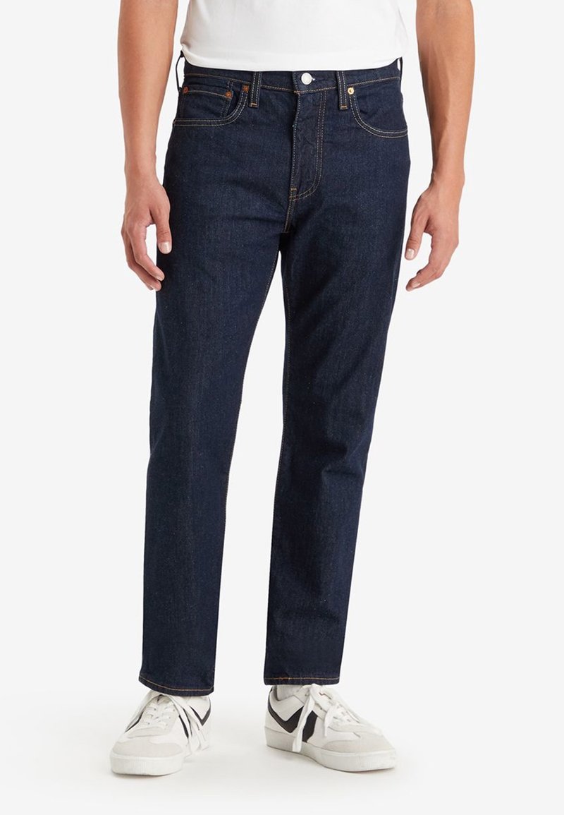 Jeans de mezclilla azul oscuro con un corte recto, que presentan remaches de cobre, costuras doradas y un diseño clásico de cinco bolsillos.