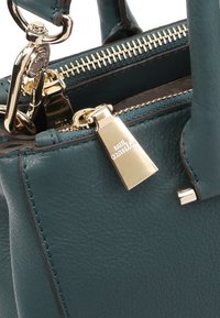 Teal leren handtas met een gestructureerd ontwerp, gouden hardware en een ritssluiting. Heeft een gemerkte metalen tag nabij de rits.