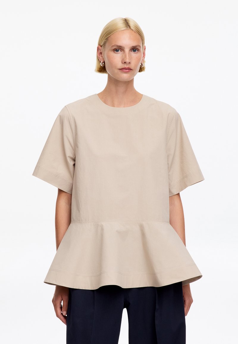 Femme blonde aux cheveux courts portant un top beige à manches courtes avec basque et un pantalon foncé sur un fond blanc uni.