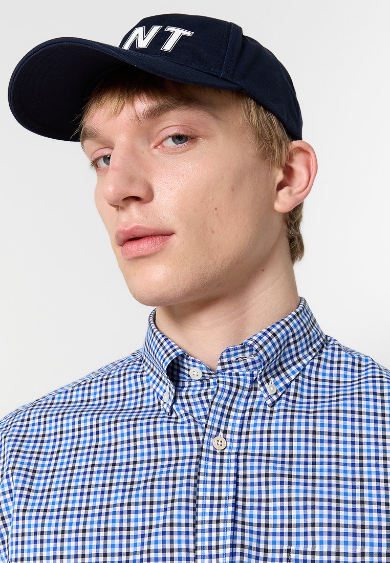 Marinkeps med vit text, blå- och vitrutigt knapp-up skjorta. Nærbild på tyget och kragadetaljer.