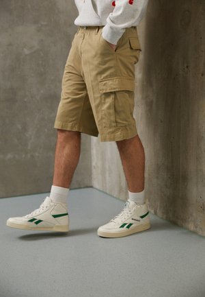 Person iført beige cargoshorts, hvide høje sneakers med grønne striber og en lys grå sweatshirt, der står ved en betonvægg.