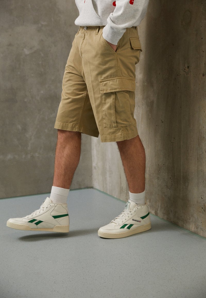 Personne portant un short cargo beige, des baskets montantes blanches avec des rayures vertes, et un sweat-shirt gris clair, debout près d'un mur en béton.