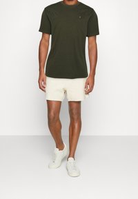 Mörkgrön t-shirt med kort ärm och rund hals, prydd med en bröstat logotyp, i kombination med krämfärgade frotté-shorts och ljusa sneakers.