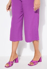 Pantalones morados cortos combinados con sandalias moradas brillantes de tacón cuadrado con un diseño de tiras cruzadas. Textura suave y una silueta moderna.