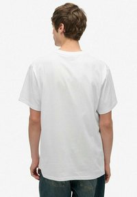 Mand med kort brunt hår iført en ensfarvet hvid t-shirt med korte ærmer og blå jeans, vendt væk fra kameraet mod en hvid baggrund.