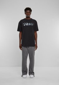 Svart oversized t-skjorte med en blå graffiti-stil grafikk, kombinert med grå vide bukser og hvite sneakers, satt mot en grå bakgrunn.