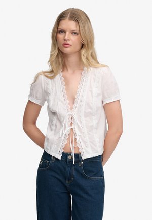 Junge Frau trägt eine weiße, kurzärmelige Bluse mit Spitzenbesatz und vorderen Bändern sowie eine hoch taillierte dunkelblaue Jeans, steht mit verschränkten Händen am Rücken.