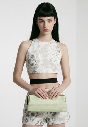 Femme portant un crop top blanc fleuri et un short assorti, tenant une pochette verte claire à motifs avec les deux mains devant sa taille.