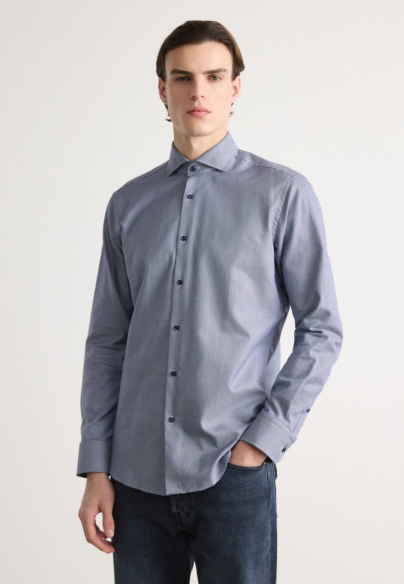 Camicia abbottonata a quadri blu e bianchi, con maniche lunghe, colletto a punta e bottoni blu. Il tessuto sembra liscio e leggero.