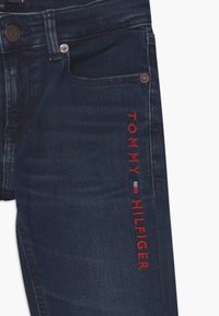 Ciemnoniebieskie dżinsy z czerwonym haftowanym napisem "TOMMY HILFIGER" z boku, z metalowymi guzikami i przednimi kieszeniami.