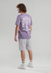 Purple cotton t-shirt with white graphic text and tree design on the back, paired with light gray shorts and white sneakers.