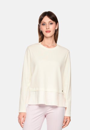 Woman with straight brown hair wears a cream long-sleeve top with a pleated hem and pale pink pants, standing against a light background.