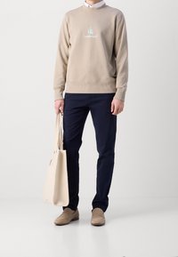 Uomo che indossa un pullover beige di Calvin Klein, pantaloni blu navy, mocassini beige, mentre tiene una borsa tote beige, in piedi contro uno sfondo bianco.