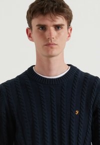Donkerblauwe gebreide trui met lange mouwen, een textuur kabelontwerp, ronde halslijn en een klein oranje geborduurd logo op de linkerkant van de borst.