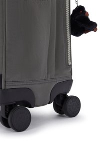 Grau farbener Stoffkoffer mit glatter Oberfläche, ausgestattet mit vier schwarzen Rollen und einem Plüschaffen als Schlüsselanhänger, der am Reißverschluss befestigt ist.