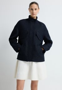 Granatowa kurtka z teksturowanej tkaniny, z kołnierzem stoj�ącym, dwiema kieszeniami z klapami oraz zamkiem błyskawicznym z przodu, w połączeniu z białą spódnicą typu dzwon.