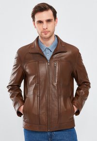 Giacca di pelle marrone con zip frontale, tasche laterali e tasca sul petto con zip. Presenta un collo a camicia e dettagli a pannello testurizzati.