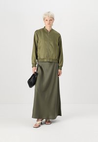 Giacca bomber verde oliva con zipper, abbinata a una lunga gonna in satin verde oliva. Una borsa nera e sandali beige completano il look.