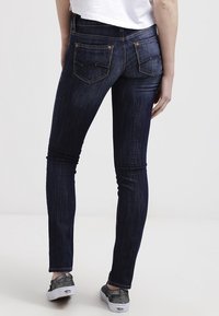 Jeans ajustados de color azul oscuro con una textura de denim suave, que cuentan con bolsillos traseros y detalles de costura sutiles, combinados con unos zapatos cómodos gris.