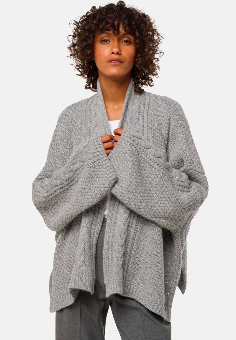 Cardigan en tricot gris avec une texture épaisse, des manches larges et des motifs en torsades sur les bras, présentant un design ouvert et confortable.