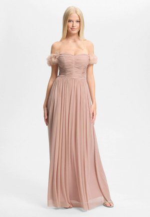 Jeune femme portant une robe longue rose poudré épaules dénudées avec un corsage froncé et des détails de fleurs en tulle sur les manches, debout devant un fond blanc.