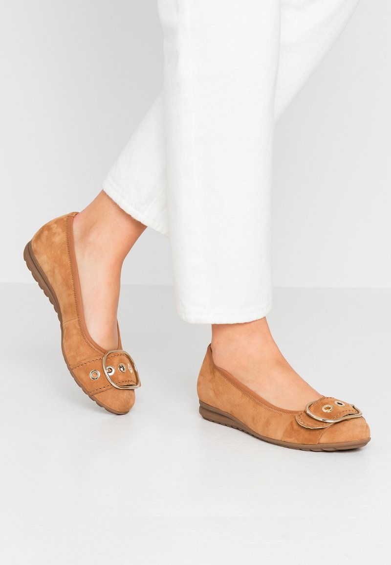 Pieds portant des chaussures plates en daim beige avec un grand détail de boucle, associées à un pantalon blanc sur un fond clair uni.