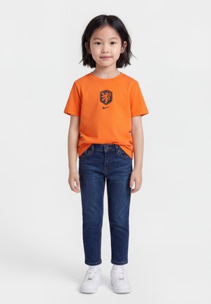 Child standing wearing an orange Nike t-shirt with a black lion crest, dark blue jeans, and white sneakers against a plain white background.