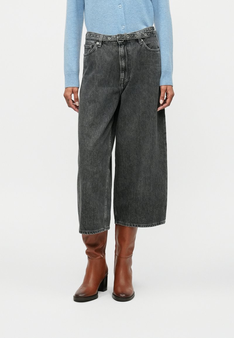 Πλατύτερο πόδι, ψηλόμεσα σκούρα γκρι τζιν κατασκευασμένα από τζιν, με θηλιές ζώνης, εμπρόσθιες τσέπες και ακατέργαστο τελείωμα. Συνδυασμένα με καφέ μποτάκια μέχρι τον αστράγαλο.