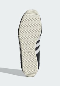 Black sneaker with textured fabric, featuring three white stripes and a cream rubber sole. The sole has a zigzag tread pattern.
