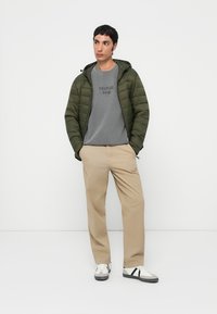 Padded green jacket, grey sweatshirt with text, beige trousers, and sporty shoes with black and white accents. Simple, functional outfit.