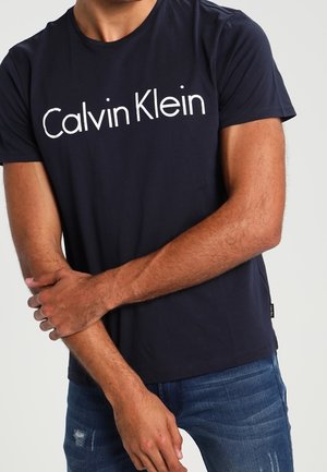 Muž v námořnicky modrém tričku Calvin Klein a roztrhaných modrých džínách, s rukama zkříženýma před sebou.