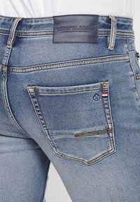 Gai zili denim džinsi ar aizmugures kabatu, kas rotāta ar daudzkrāsainām svītrām, oranžu šuvju un tumši zilu ādas zīmolu tagu jostasvietā.