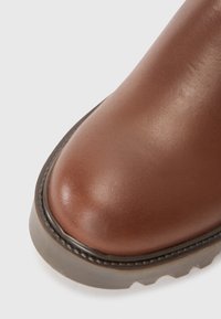 Bota de cuero marrón con una textura suave, punta redonda y una suela de goma gruesa con un patrón en zigzag. Detalles de costura a lo largo del borde.