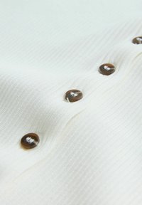 Närbild av vit ribbad tyg med en rad av fyra bruna knappar sydda med vit tråd.