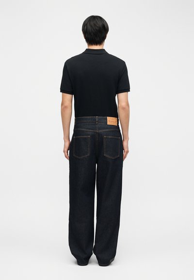 Άτομο που φοράει μαύρο πόλο με κοντό μανίκι και φαρδιά σκουρόμπλε τζιν, στέκεται γυρισμένο με την πλάτη σε απλό λευκό φόντο.