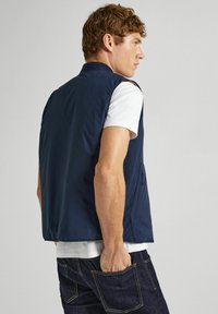 Marinblå ärmlös väst i slät tyg, med hög krage, sidofickor och avslappnad passform, bärs över en vit t-shirt.