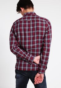 Camicia a quadri rossa e grigia con maniche lunghe, colletto button-down e dettaglio sul yoke posteriore. Il materiale sembra morbido e strutturato.