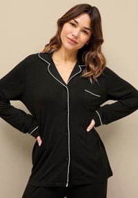 Sort pyjamas top med lange ærmer, hvid piping langs kraven og lommen, knaplukning og en enkelt brystlomme. Blødt materiale.