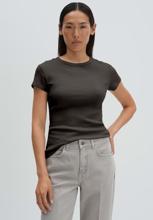 Vrouw met steil donker haar die een aansluitend zwart T-shirt met korte mouwen en lichtgrijze broek met hoge taille draagt, staand tegen een effen achtergrond.