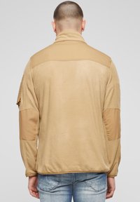 Beige Fleecejacke mit hohem Kragen, kontrastierenden Schulter- und Ärmelpartien. Glatte Textur mit lockerem Schnitt und minimalen Details.