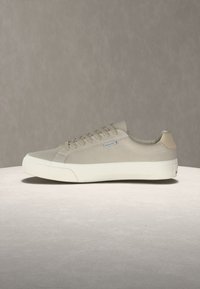 Χαμηλό μπεζ αθλητικό παπούτσι με λευκή παχιά σόλα, σχέδιο με κορδόνια και μικρή μπλε ετικέτα στο πλάι, εμφανίζεται σε ουδέτερο φόντο.