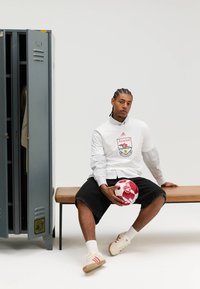 Witte lange mouwen blouse over gestreept overhemd met kraag, zwarte korte broek, zittend op een bankje met een rood-witte voetbal naast een grijze kluis.