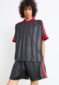 Giovane con capelli ricci che indossa un completo sportivo Nike nero e rosso, in piedi davanti a uno sfondo bianco uniforme.
