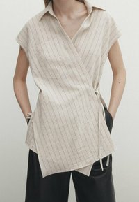 Top senza maniche a righe beige realizzato in un tessuto leggero, con colletto e dettagli con fiocco laterale. Presenta una tasca sul petto e una vestibilità ampia.
