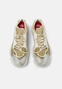 Beige athletic sneakers featuring a mesh upper, curved sole, white laces, and layered texture with a wavy pattern in cream and brown.