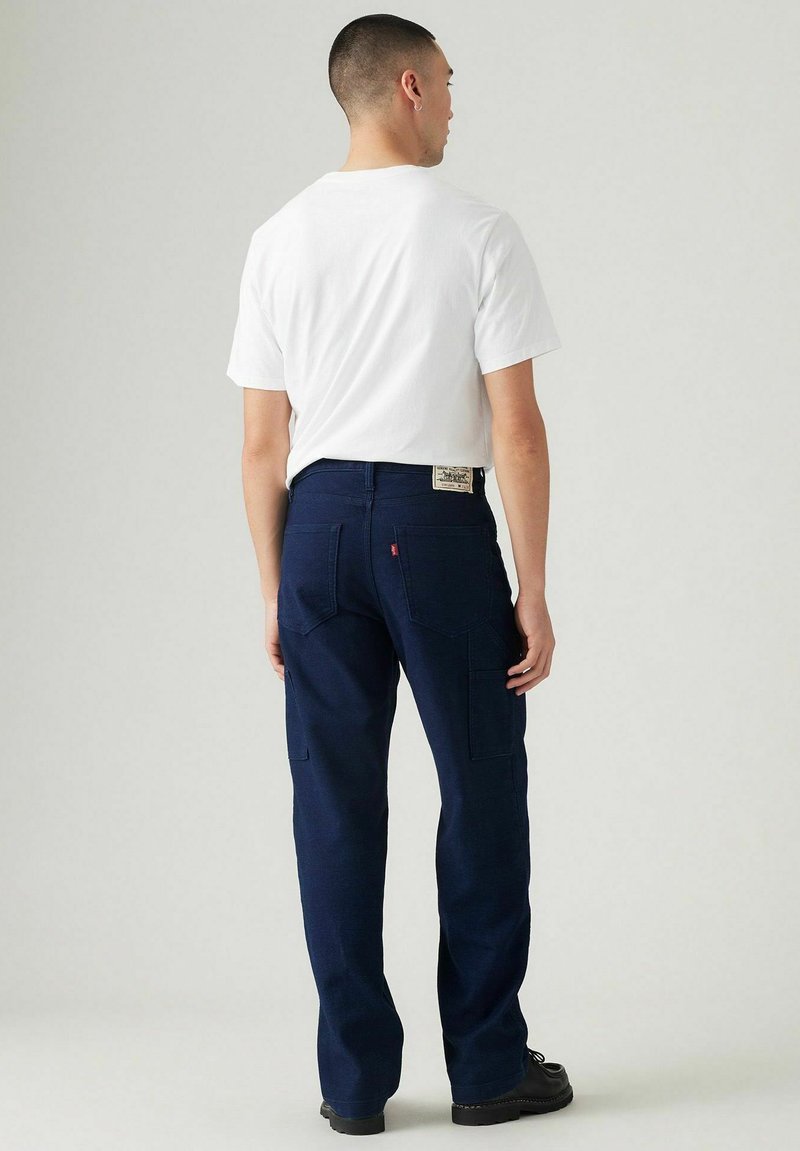Pantalones cargo azul marino en un diseño de pierna recta con bolsillos traseros y una etiqueta sutil. Combinados con una camiseta blanca y zapatos negros.